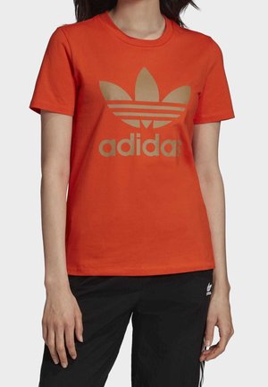 Kobieta w pomarańczowej koszulce Adidas z brązowym logo i czarnych spodniach Adidas, stojąca na jednolitym jasnym tle.