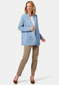 Helles blaues Cardigan aus weichem Stoff, mit einem Schalkragen, zwei Fronttaschen und gerippten Bündchen. Kombiniert mit beigen Hosen und braunen High Heels.