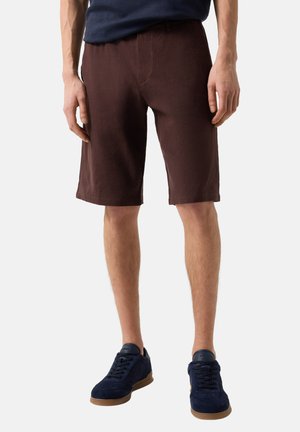 Mann trägt dunkelbraune kniehohe Shorts, marineblaue Sneakers mit Kautschuksohlen und ein marineblaues Hemd, steht mit entspannten Armen.