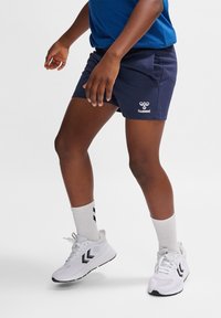 Marinblå shorts med sidofickor, prydda med en vit logotyp. Vida sportskor i vitt med svarta detaljer och ljusgrå ankelsockor med svarta ränder.