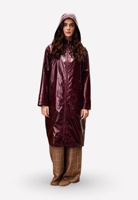 Bordeaux glanzende regenjas met capuchon, volledige rits aan de voorkant en lange mouwen, gecombineerd met gemusterde bruine wijde broek en tan schoenen.