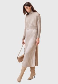 Maglione beige a collo alto abbinato a una gonna plissettata lunga fino alla caviglia in tonalità simile. Completano l'outfit stivali beige alti fino al ginocchio con punta. 