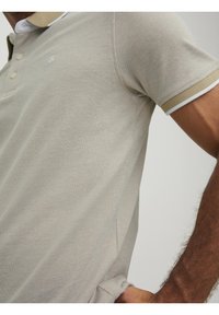 Jasno beżowa koszulka polo o teksturowanej powierzchni, z kontrastowym kołnierzykiem i mankietami w ciemniejszym odcieniu oraz z trzema guzikami.