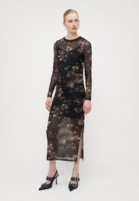 Robe longue en voilage noir à manches longues avec fentes sur les côtés, dotée d'un corsage ajusté et d'une jupe maxi fluide. Chaussures à talons hauts noires avec boucles.