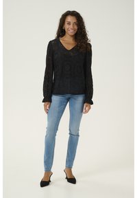 Top lungo in pizzo nero con maniche lunghe, scollo a V e motivi testurizzati, abbinato a jeans skinny blu chiaro e tacchi a punta neri.