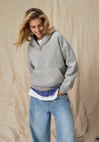 Szara bluza z kapturem na zamek, wykonana z miękkiego materiału, z przednią kieszenią i ściągaczem u dołu, noszona na białej koszuli w niebieskie paski i jasnych dżinsach.