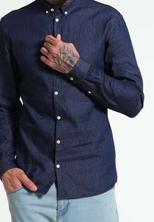 Mann trägt ein dunkelblaues, geknöpftes Jeanshemd mit hellblauer Jeans, zeigt eine tätowierte Hand, die auf der Brust ruht.