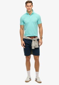 Polo azzurro chiaro, pantaloni corti blu navy scuri, maglione grigio annodato in vita, cintura marrone, calzini bianchi e scarpe da ginnastica con dettagli grigi.