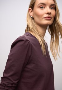 Dunkellila Langarmoberteil mit rundem Ausschnitt und leichtem Puffärmel-Detail an den Schultern. Weicher, figurbetonter Stoff. Model mit langen blonden Haaren.