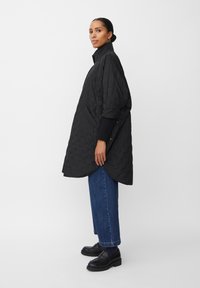 Svart vadderad kappa med en strukturerad krage, stor silhuett, sidofickor och subtilt mönster, matchad med blå jeans och svarta skor.