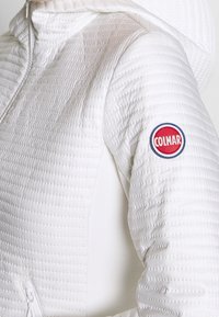 Vit pufferjacka med ett quiltat mönster, hög krage, sidofickor med blixtlås och en cirkulär röd, vit och blå logotyp på axeln.