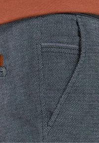 Gros plan sur la poche d'un pantalon en tissu gris texturé avec une petite poche fendue au-dessus, porté avec un haut orange.
