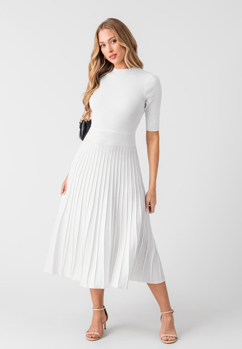 Robe blanche plissée avec des manches courtes, texture côtelée, corsage ajusté et jupe évasée. Complétée par des talons nus à lanières.