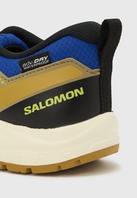 Κοντινό πλάνο της φτέρνας αδιάβροχου παπουτσιού Salomon σε μπλε, μαύρο και καφέ χρώμα με λευκή σόλα και μαύρο λουράκι έλξης.