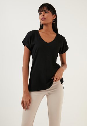 Black short-sleeve top with a V-neck, textured fabric, relaxed fit, and curved hem. Paired with beige slim-fit trousers.