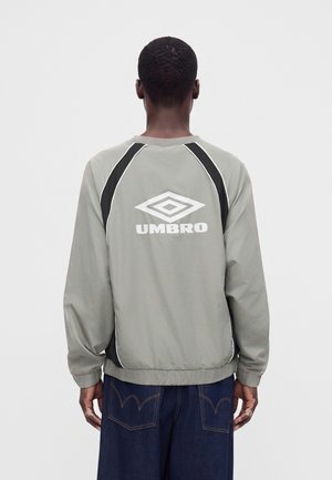 Άτομο φορώντας γκρι και μαύρο μπουφάν Umbro με λογότυπο στην πλάτη, συνδυασμένο με σκούρο μπλε τζιν, φαίνεται από πίσω.