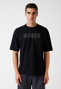 Must have mustamust ja lühikeste varrukatega T-särk, mille keskosas on silmapaistvalt väljaulatuv hall "GUESS" logo ja tavaline ümar kaelus.