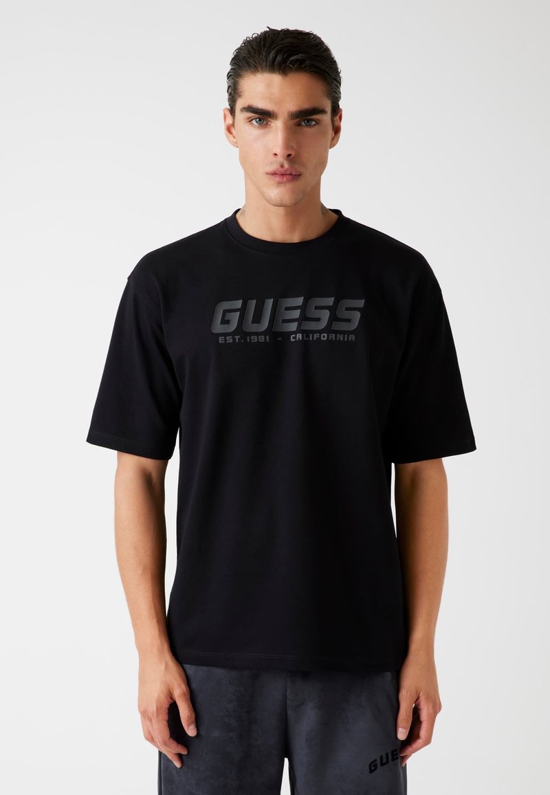 Must have mustamust ja lühikeste varrukatega T-särk, mille keskosas on silmapaistvalt väljaulatuv hall "GUESS" logo ja tavaline ümar kaelus.