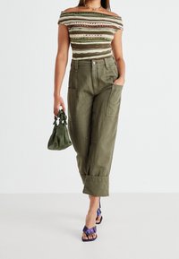 Top off-the-shoulder con rayas multicolores, combinado con pantalones cargo de pierna ancha en verde oliva y un pequeño bolso verde. Sandalias de tacón alto en púrpura.