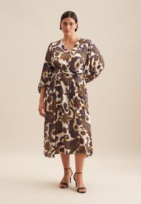 Langes, gemustertes Kleid mit V-Ausschnitt, Puffärmeln und gebundenem Taillenbund in Creme-, Blau- und Goldtönen. Getragen mit schwarzen hohen Schuhen.