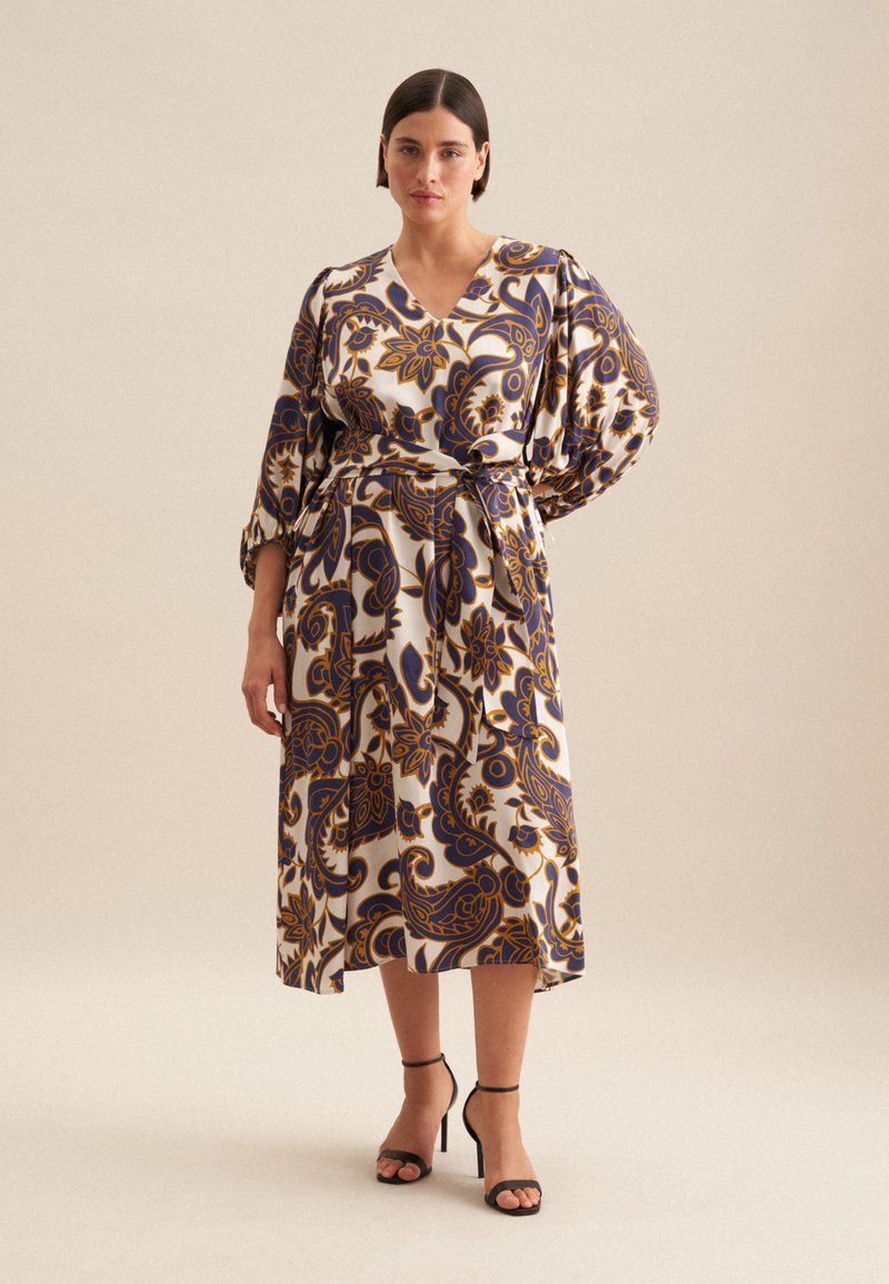 Langes, gemustertes Kleid mit V-Ausschnitt, Puffärmeln und gebundenem Taillenbund in Creme-, Blau- und Goldtönen. Getragen mit schwarzen hohen Schuhen.