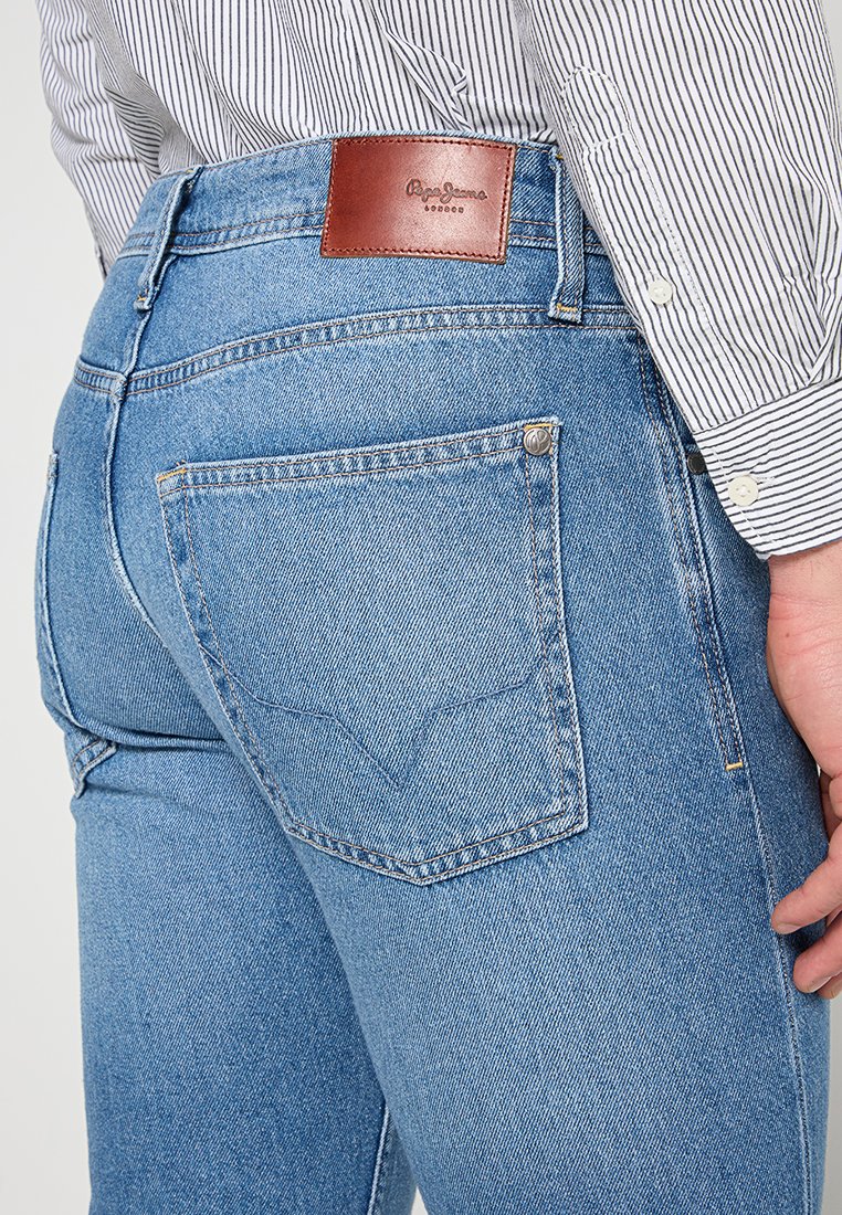 Primer plano de una persona que lleva vaqueros de denim de Pepe Jeans en azul claro y una camisa de manga larga blanca a rayas, mostrando el bolsillo trasero y el parche de cuero de la marca.