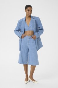 Tenue en denim composée d'un top court et de shorts à jambes larges en bleu clair, agrémentée d'un blazer ample. Talons blancs avec accents ronds.