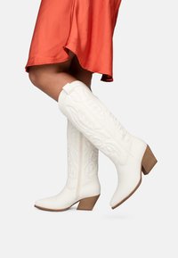 Botas blancas hasta la rodilla, hechas de material texturizado, con un diseño ornamentado, punta afilada y un tacón de madera apilada.
