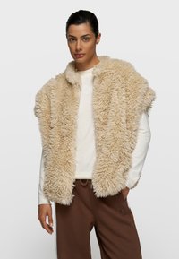 Chaleco de piel sintética beige con un acabado suave y texturizado; diseño de frente abierto; llevado sobre una camisa de manga larga de color claro y pantalones marrones.