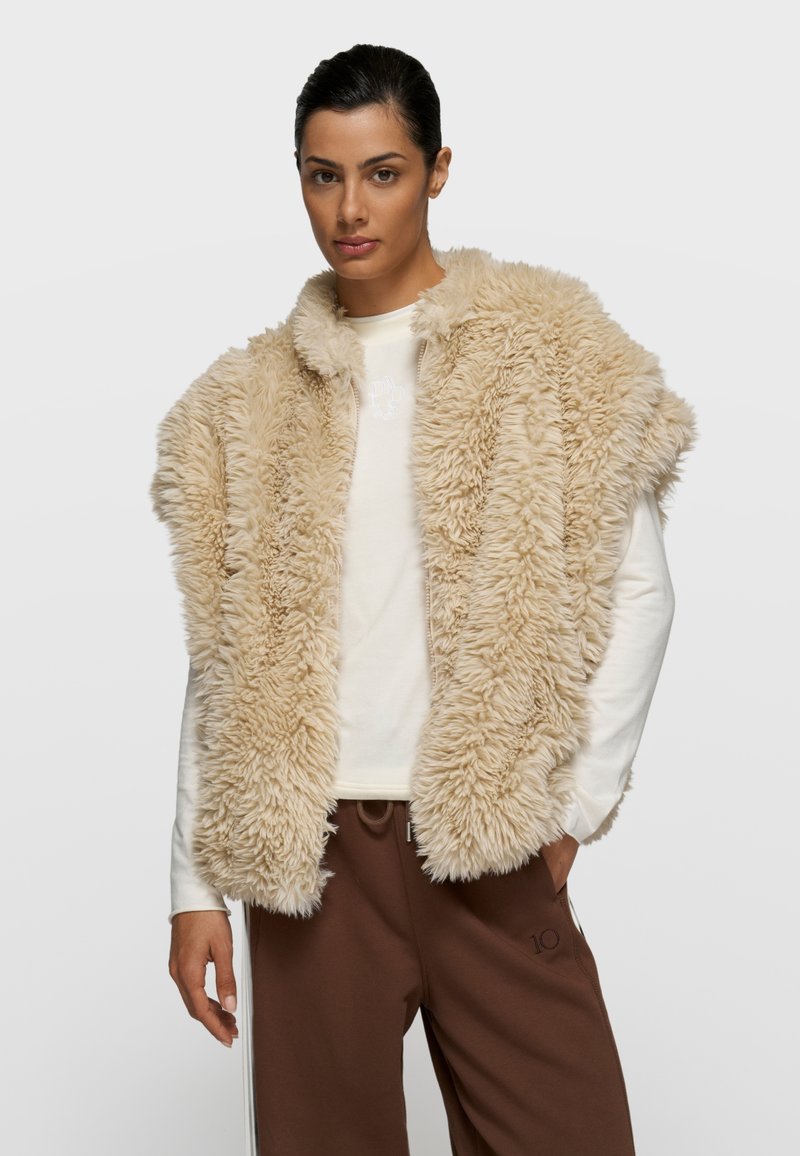 Chaleco de piel sintética beige con un acabado suave y texturizado; diseño de frente abierto; llevado sobre una camisa de manga larga de color claro y pantalones marrones.