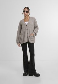 Bež pleteni cardigan z V-izrezom, sprednjim zapiranjem na gumbe in dvema velikima žepoma. Nosi se čez belo majico in je kombiniran z razširjenimi črnimi hlačami.