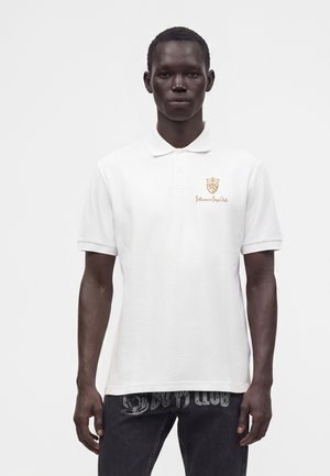 Homme portant un polo blanc avec un petit logo doré "Billionaire Boys Club" sur la poitrine gauche, associé à un jean noir.