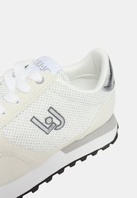 Scarpe da ginnastica bianche in mesh con una tomaia liscia e testurizzata, dettagli in argento, colletto imbottito e suola in gomma nera. Logo sul lato.