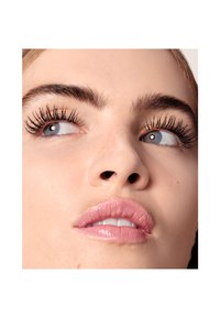 Nahaufnahme eines Gesichts mit ausgeprägten, definierten Wimpern, glatter Haut und vollen Lippen in einem hellen Rosaton. Die Augenbrauen sind gepflegt und dunkel.