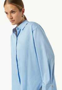 Light blue button-up shirt with a relaxed fit, pointed collar, and oversized sleeves. Features a side slit and decorative button accents.