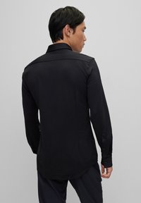 Zwarte langemouwen shirt met een klassieke kraag, een gladde textuur en een gebogen zoom. Knopenmanchetten en een getailleerde pasvorm aan de achterkant.
