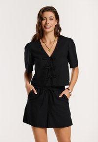 Top negru, cu mânecă scurtă și decolteu în v, având un fâșie la partea din față. Asortat cu pantaloni scurți negri, ambele dintr-un material ușor, prezentând o textură subtilă.