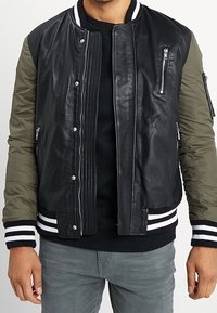 Blouson aviateur pour homme avec un corps en cuir noir, des manches vert olive, des poignets côtelés rayés blancs et noirs, et une poche poitrine zippée.