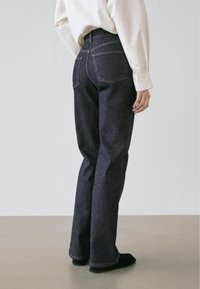 Jeans in denim blu scuro con vita alta, gamba a zampa e due tasche posteriori ben visibili. Indossati con una camicia bianca a maniche lunghe e scarpe nere.