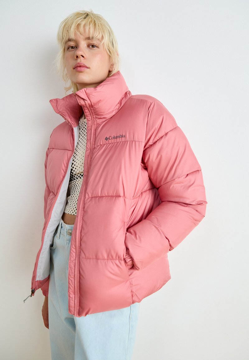 Doudoune rose avec un col haut, présentant une texture lisse, un matelassage horizontal et un logo Columbia sur la poitrine gauche.