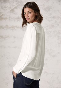 Witte blouse met lange mouwen, gemaakt van een lichtgewicht stof, met een rugplooi detail, in combinatie met donkerblauwe jeans en een textuur achtergrond.