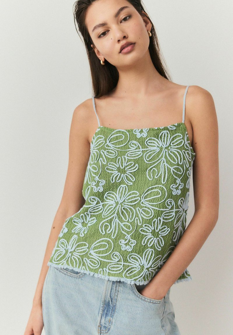 Top verde brodat cu modele florale albe și albastre, cu bretele subțiri. Material texturat cu margine franjurată, asociat cu pantaloni scurți din denim.