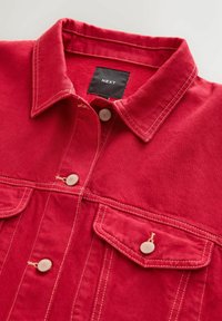 Red denim jacket with a pointed collar, two front pockets, and beige stitching. Features round buttons and a black brand label.