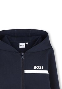 Sweat à capuche zippé bleu marine avec capuche, affichant le logo "BOSS" blanc et une bande blanche horizontale traversant la poitrine.