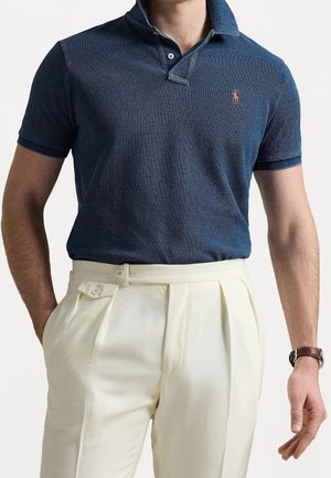 Hombre vistiendo una camiseta tipo polo de manga corta azul marino con logo rojo y pantalones plisados crema de talle alto, con una mano en el bolsillo y un reloj marrón en la muñeca.