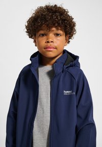 Mörkblå zip-up jacka med huva, med svart insida, bärs över en ljusgrå skjorta. Logotyp på vänster bröstområde. Mjuk tygtextur.
