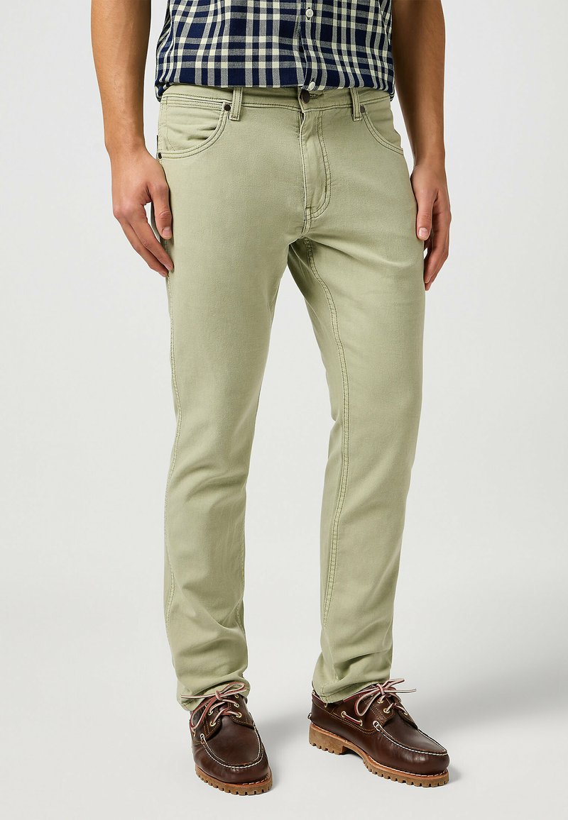 Vaqueros de corte ajustado en verde claro con una textura suave, cinco bolsillos y costuras en contraste. Combinados con zapatos de barco marrones.