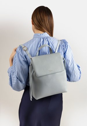 Donna con capelli castani che indossa una camicetta a righe blu e una gonna blu scuro porta uno zaino in pelle grigio chiaro sulla schiena su uno sfondo semplice.