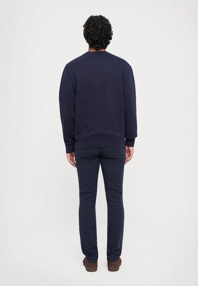 Felpa girocollo blu marina con polsini a coste, abbinata a pantaloni slim-fit blu marina e scarpe marroni, vista da dietro su uno sfondo neutro.