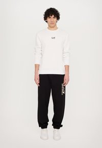 White crew neck sweatshirt with black EA7 logo, paired with black sweatpants featuring a gold logo print. White sneakers complete the look.